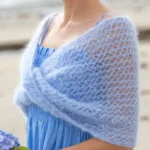 Soft blue mohair infinity wrap worn over a light blue dress, featuring an airy open-knit texture and a twisted front detail, styled as an elegant shoulder cover for special occasions.