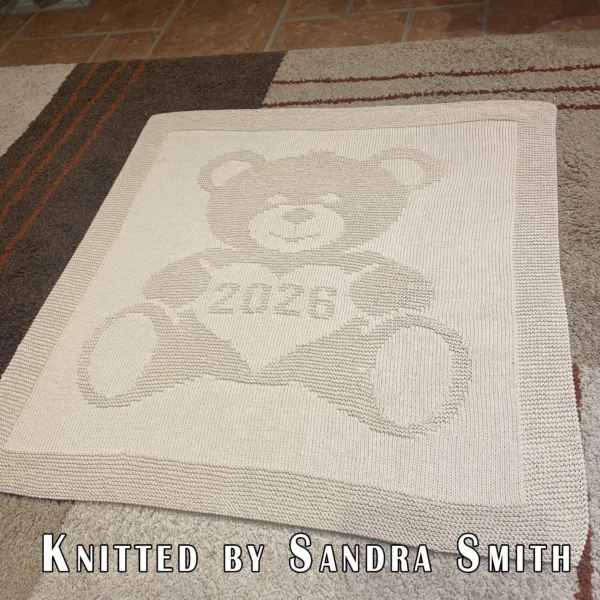Beige knitted baby blanket featuring a textured teddy bear holding a heart with “2026” inside. The teddy bear and border are worked in raised garter stitch against a smooth stockinette background, creating a soft embossed effect. Handmade-style nursery blanket design for newborn babies and baby shower gifts.