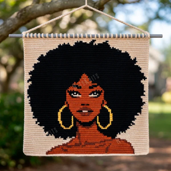 Tapestry crochet graphghan panel showing a portrait of a woman with a large natural afro, warm caramel and brandy skin tones, gold hoop earrings and an ivory background, worked in single crochet intarsia colorwork with 5 colors: Black, Ivory, Caramel, Brandy, and Gold.