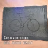 Knitted blanket featuring a bicycle motif in the center, worked in garter stitch against a stockinette background. The blanket has a textured border framing the bike design. The bicycle is shown in a side profile with detailed wheels, frame, handlebars, and pedals, suitable for cyclists, bike lovers, teens, and adults.