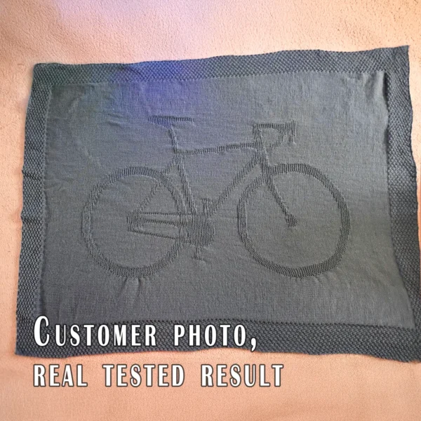 Knitted blanket featuring a bicycle motif in the center, worked in garter stitch against a stockinette background. The blanket has a textured border framing the bike design. The bicycle is shown in a side profile with detailed wheels, frame, handlebars, and pedals, suitable for cyclists, bike lovers, teens, and adults.