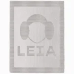 Minimalist knitted blanket featuring a stylized Princess Leia face silhouette with her iconic side buns and the word “LEIA” below. The design is worked in textured garter stitch against a smooth stockinette background, all in a soft neutral color palette with a wide garter stitch border. A modern and cozy blanket design inspired by classic sci-fi themes, perfect for kids’ rooms, reading nooks, movie nights, and Star Wars fans of all ages.