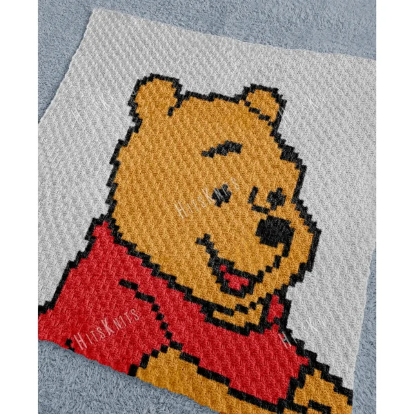 Pixel-style Winnie the Pooh C2C crochet blanket in soft plush yarn, featuring Pooh Bear wearing a red shirt on a light gray background. Handmade graphghan blanket with cartoon-inspired design and cozy textured stitches.
