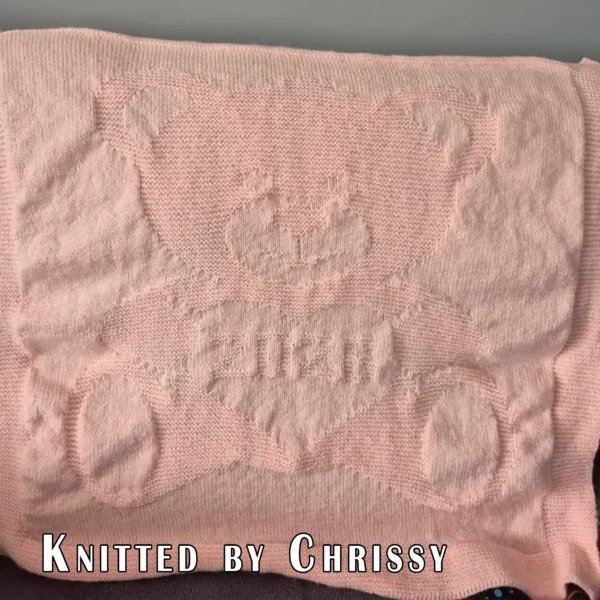 Beige knitted baby blanket featuring a textured teddy bear holding a heart with “2026” inside. The teddy bear and border are worked in raised garter stitch against a smooth stockinette background, creating a soft embossed effect. Handmade-style nursery blanket design for newborn babies and baby shower gifts.