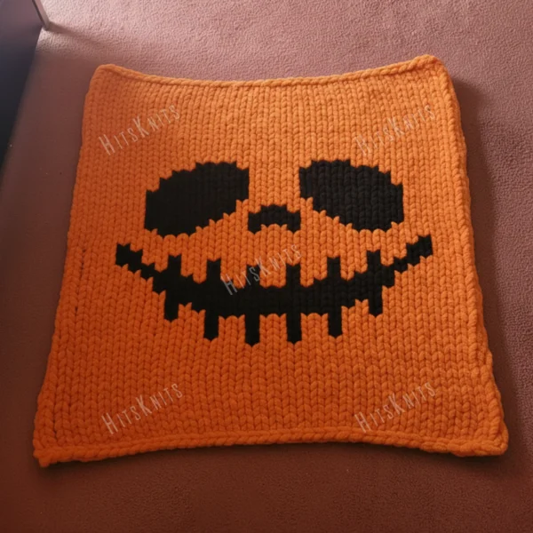 Jack Skellington Finger Knitting Pattern - a grinning skeleton face with large slanted oval eyes, a small triangular nose and a wide stitched smile on a bright orange background, knitted in stockinette colorwork intarsia with 2 colors: Orange, Black.