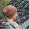 Machine Knitting Slouchy Hat Pattern showing a woman in profile wearing a warm brown cotton beanie with a mock rib cuff, worn slouchy, styled with a floral scarf and a light jacket against a blurred autumn landscape.