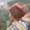 Machine Knitting Slouchy Hat Pattern showing a woman in profile wearing a warm brown cotton beanie with a mock rib cuff, worn slouchy, styled with a floral scarf and a light jacket against a blurred autumn landscape.
