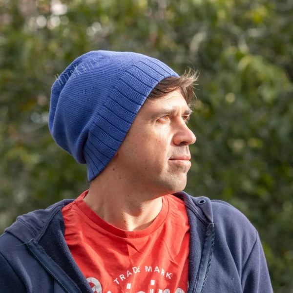 Machine Knitting Slouchy Hat Pattern showing a man wearing a royal blue cotton beanie with a mock rib cuff, worn slouchy, with a navy hoodie and a red t-shirt against a sunny green garden background.