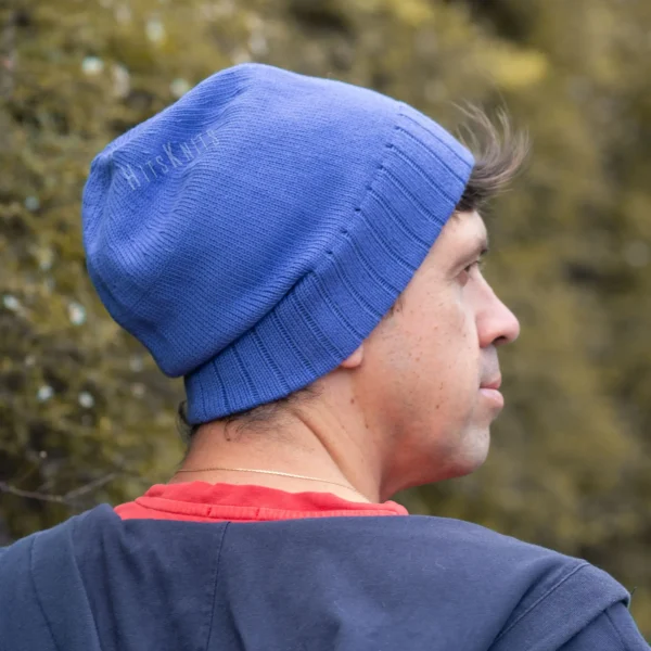 Machine Knitting Slouchy Hat Pattern showing a man from behind in profile wearing a royal blue cotton beanie with the mock rib cuff turned up, styled with a navy hoodie and a red t-shirt against a blurred green background.