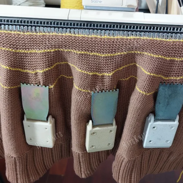 Machine Knitting Slouchy Hat Pattern showing the brown cotton hat in progress on a Brother standard gauge knitting machine, with claw weights hung evenly on the fabric, the mock rib cuff at the bottom and yellow waste yarn rows marking the crown decreases.