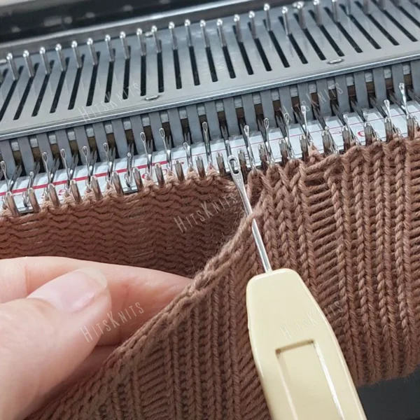 Machine Knitting Slouchy Hat Pattern showing the brown cotton hat in progress on a Brother standard gauge knitting machine, with claw weights hung evenly on the fabric, the mock rib cuff at the bottom and yellow waste yarn rows marking the crown decreases.