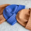 Machine Knitting Slouchy Hat Pattern showing finished cotton beanies in warm brown and royal blue with a mock rib cuff, laid flat and stacked to show the smooth stockinette body, the round crown and the cuff worn down or turned up.