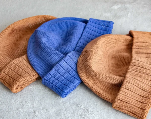 Machine Knitting Slouchy Hat Pattern showing finished cotton beanies in warm brown and royal blue with a mock rib cuff, laid flat and stacked to show the smooth stockinette body, the round crown and the cuff worn down or turned up.